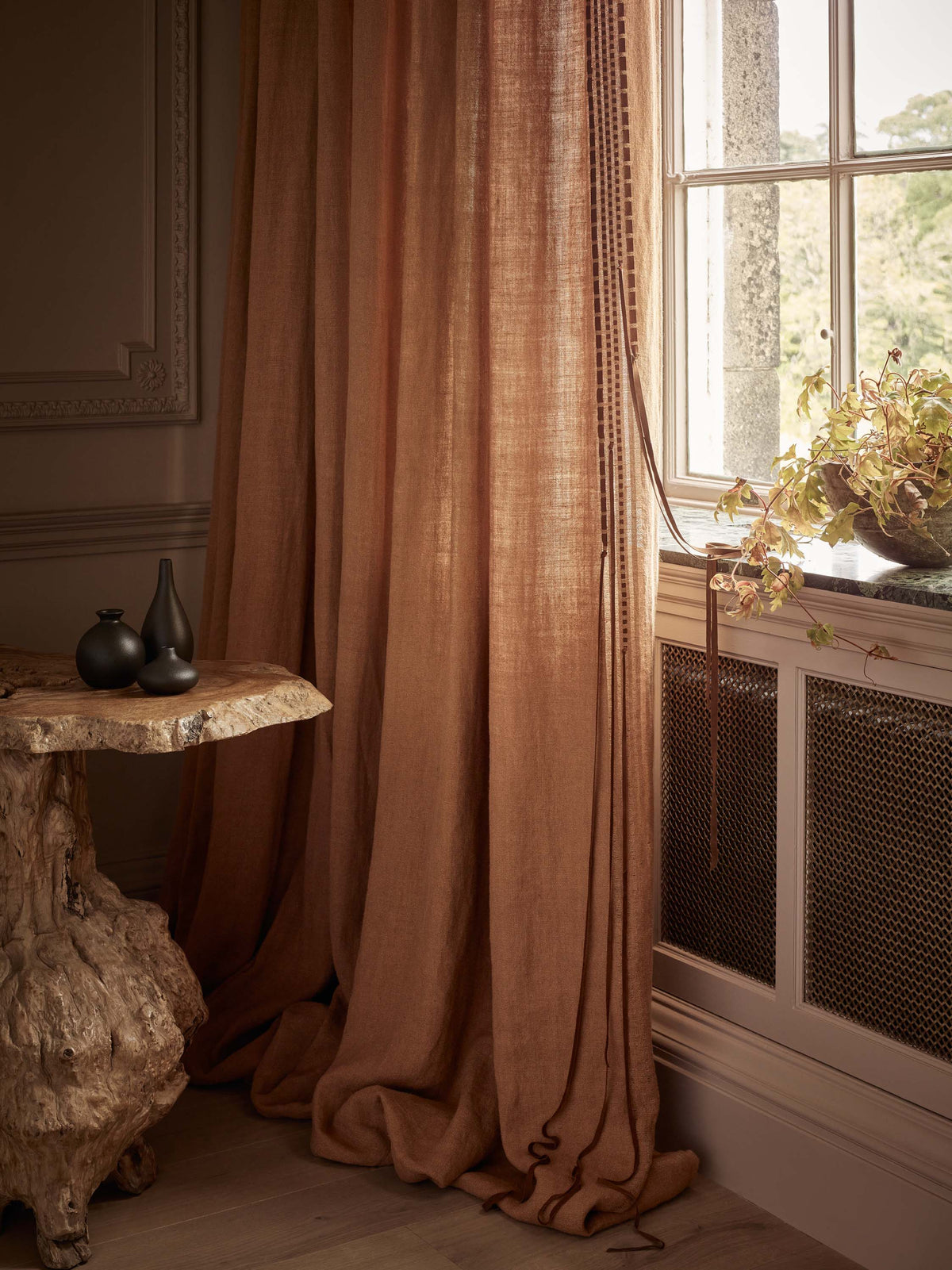 Floor-to-ceiling brown linen curtains displayed beside a large window, with natural light highlighting the fabric's texture and drape. A rustic wooden table and decorative accessories complete the interior scene.