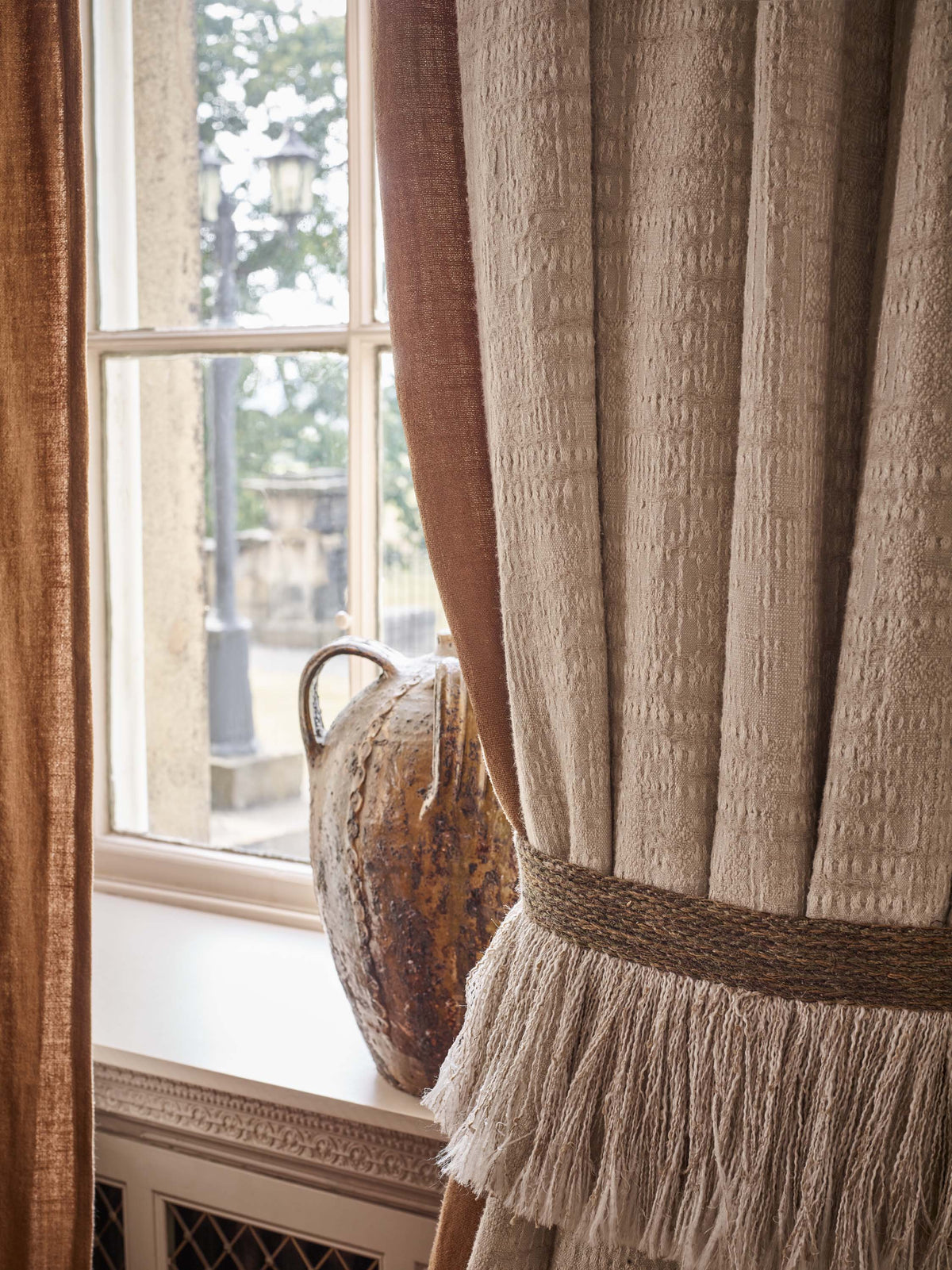 Neutral-toned textured curtains tied back with a woven fringed tieback beside a large window. Natural light illuminates the fabric's texture, while a ceramic vessel is displayed on the windowsill.
