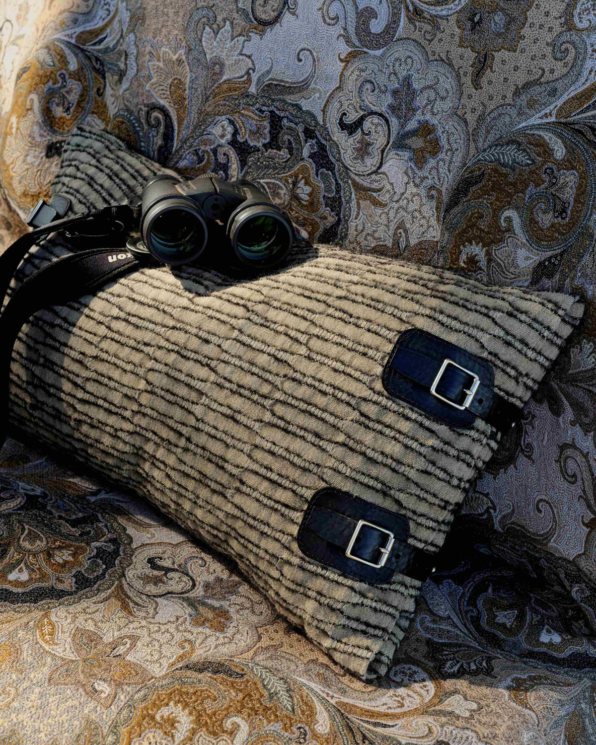 A striped beige and black travel pillow with two black buckle straps, styled on a patterned paisley sofa with a pair of black binoculars resting on top.