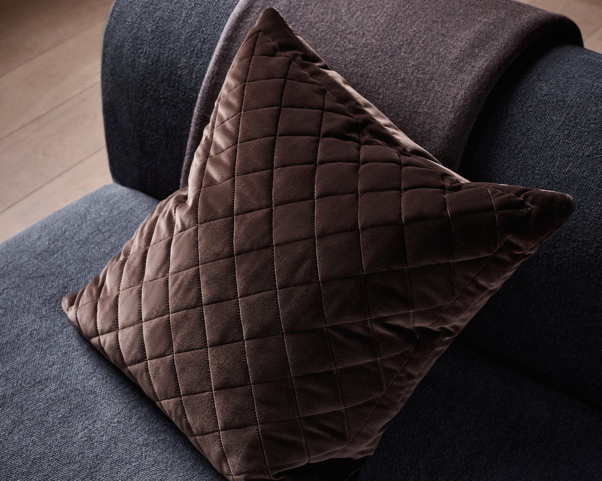 A dark brown quilted velvet throw pillow resting on a dark gray sofa with a matching brown throw blanket draped over the arm, set against a wooden floor background.