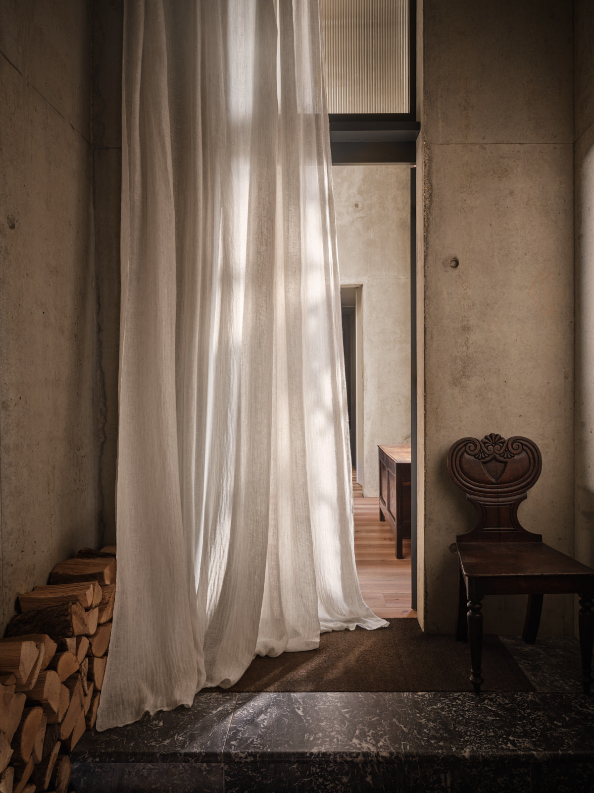 Light filtering through sheer curtains in a room with wooden floor and stacked logs.